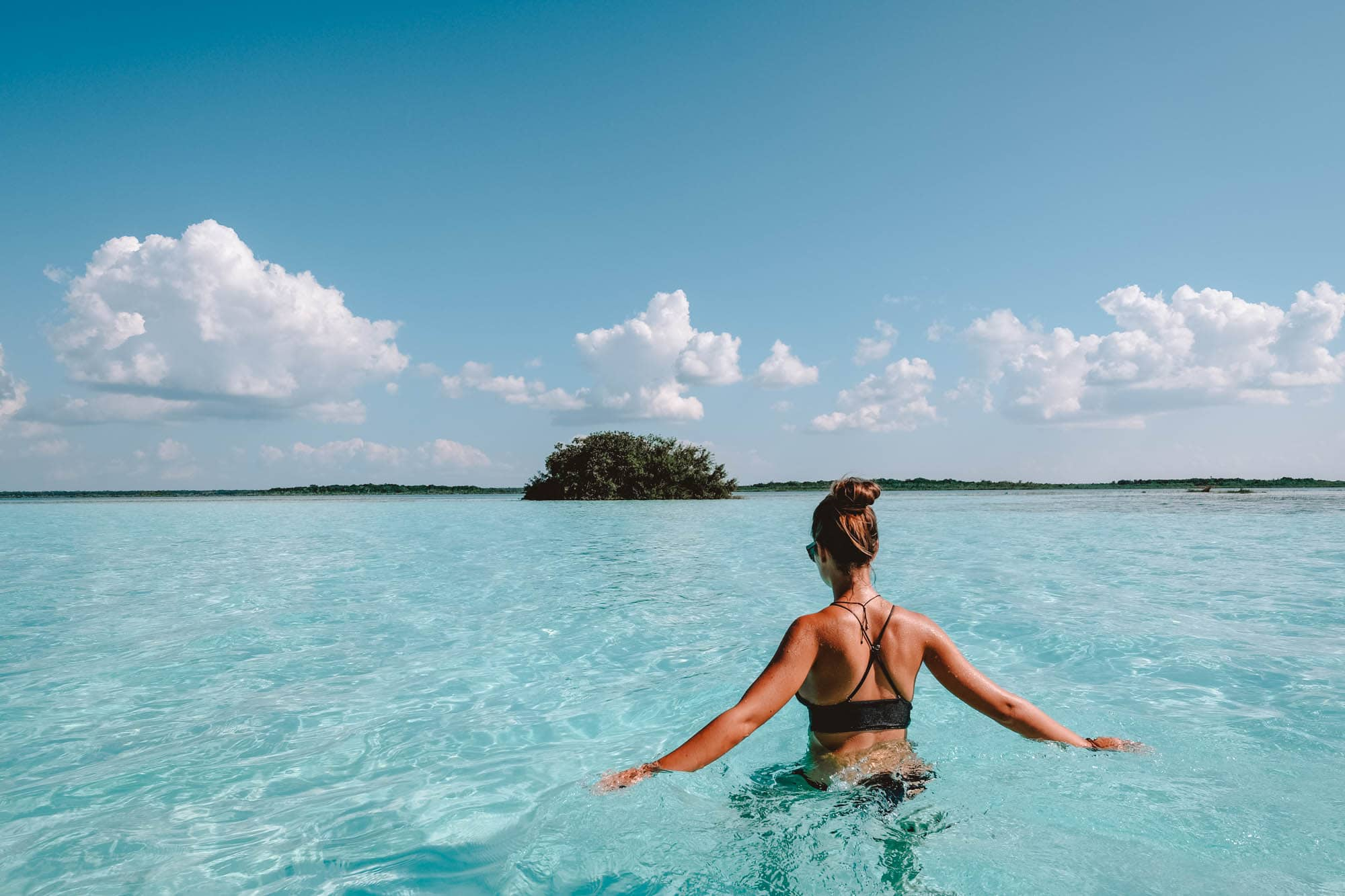 Mujer en bikini negro en agua turquesa, de espaldas, mirando hacia isla lejana. Cielo azul con nubes, ambiente sereno y relajante.